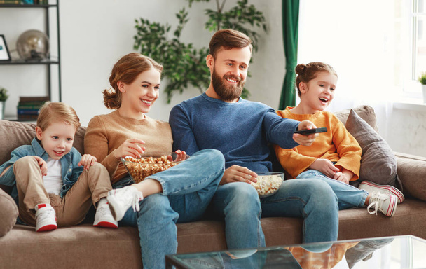 Счастливые молодые родители с симпатичными детьми, сидящими дома на диване в гостиной и смотрящими забавный фильм, поедая кукурузу и наслаждаясь выходными.
 - Фото, изображение