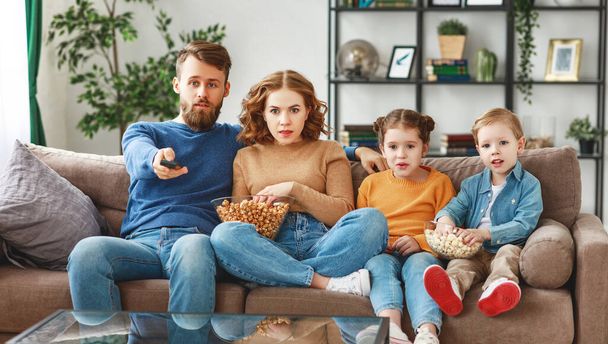 Skoncentrowany rodzice w casual ubrania siedzi na dużej kanapie z uroczymi dziećmi i oglądania ciekawych ekscytujących filmów podczas weekendu w przytulnym hom - Zdjęcie, obraz