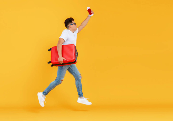 Seitenansicht eines jungen Mannes mit Koffer, der aufspringt und vor gelbem Hintergrund ein Selfie macht, während er sich auf den Sommerurlaub vorbereitet - Foto, Bild