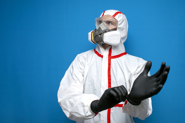Epidemiologist in a chemical protective suit puts on gloves on a blue isolated background, man, biologist, scientist, virologist looks at copyspace - Photo, Image