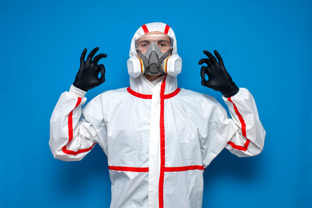 man in chemical protective suit shows okay sign on a blue isolated background, epidemiologist, biologist, virologist, scientist - Фото, зображення