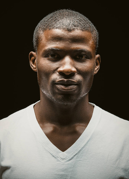 Confident Afro American man thoughtfully looking at camera. Serious attractive dark-skinned guy in a white t-shirt on black background. Toned image - Foto, Imagem