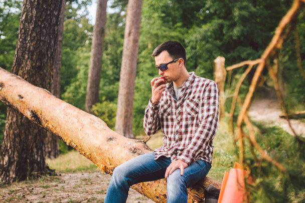 Mies syö omenaa patikoidessaan metsässä. Patikoija pitää taukoa vaeltaessaan erämaassa. Mies syö omenaa kaatuneessa puussa lähellä järveä. Helathy lifestyle, terveellinen syöminen
. - Valokuva, kuva