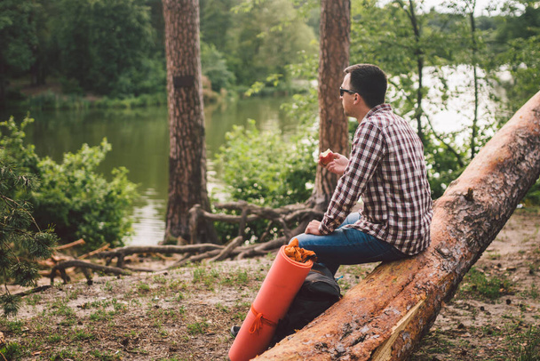 Het thema is gezond eten en vegetarisme. Buitenactiviteiten in het bos en fruitsnack. Jonge blanke mannelijke wandelaar rust en eet een appel op een open plek in de buurt van een bos meer. - Foto, afbeelding