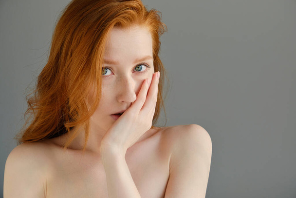 Head shot. Red girl with bare shoulders stands on a gray background, looking into the frame, covering part of the face with hand - Fotoğraf, Görsel