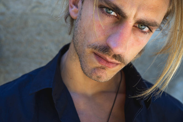 Joven modelo masculino con pelo rubio medio largo, con un collar y ojos azules, vestido con una camisa y mirando directamente a la cámara
. - Foto, imagen