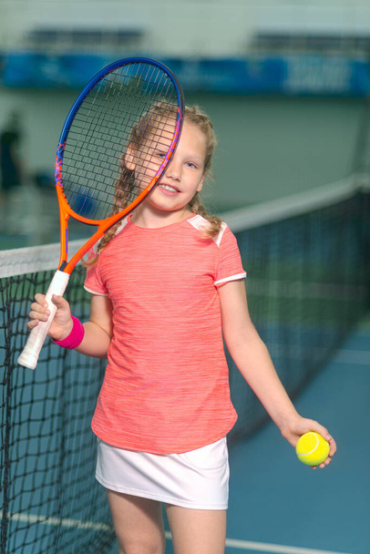 A happy playful girl smiles on the tennis court with a racket. Little girl with tennis racket and ball in sport club. Active exercise for kids. Training for young kid. Child learning to play. - Foto, Imagen
