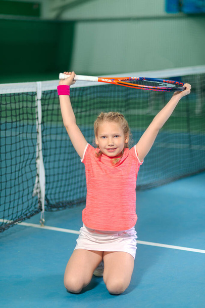A little girl on an indoor tennis court is happy that she just won a tennis match. Little girl with tennis racket and ball in sport club. Training for young kid. Child learning to play. - Photo, Image