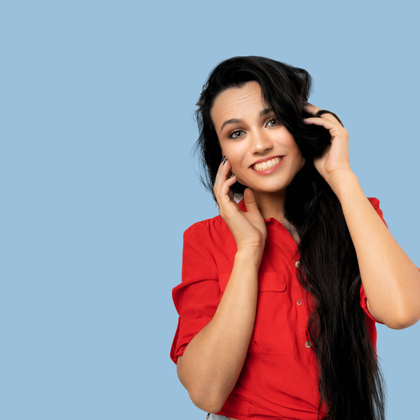 Positive young woman smiling broadly while touching face with hands. Cute long-haired brunette in a red blouse smiling while looking at the camera on a colored background. Beauty concept - Foto, Imagen