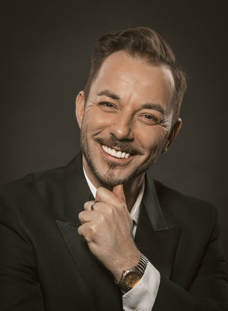 Well-dressed businessman toothy smiles while touching his chin with his hand. Portrait of handsome caucasian man in black suit on dark background. Successful business concept - Photo, Image