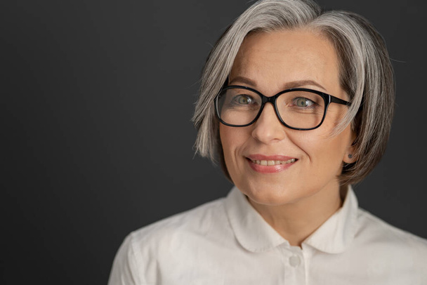 Pretty gray-haired woman in white shirt. intelligent middle-aged businesswoman in glasses with medium length care hairstyle smiling on grey background. Close up portrait. Textspace at left side - Фото, изображение