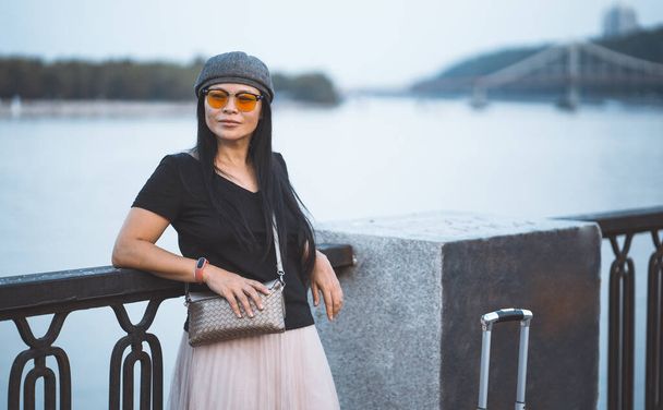 Female traveler in sunglasses resting on embankment of the river. Beautiful asian woman with suitcase travels in european city. Travel concept - Φωτογραφία, εικόνα