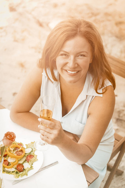 Smiling woman holds glass of white wine sitting at table On the sandy beach outdoors. Happy middle aged woman have date near the sea. High angle view. Toned image - Foto, Imagen