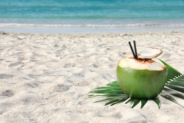 Green coconut with refreshing drink and palm leaf on sandy beach near sea, space for text - Φωτογραφία, εικόνα