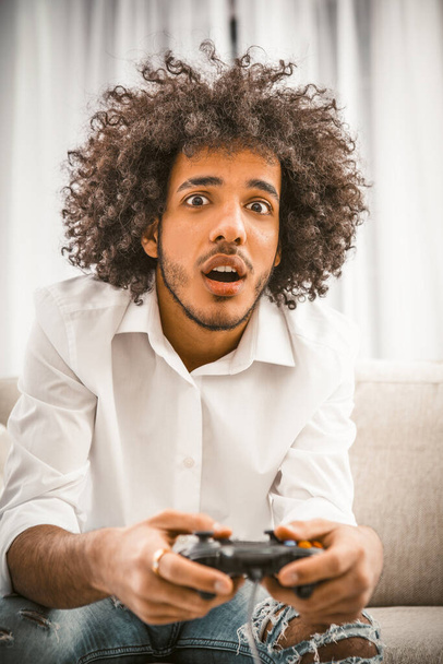 Gamer játszani számítógépes játék otthon elszigetelten. Meglep, hogy Arab Fiatal Meleg tartja Play Station joystick ül kanapén. Számítógépes játék koncepció. Karantén koncepció. Tonozott kép - Fotó, kép