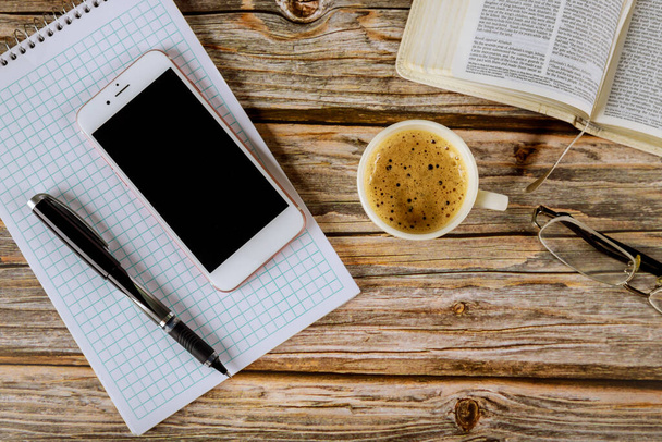 Λος Άντζελες CA US 16 MAY 2020: Μελέτη με Αγία Γραφή με μαύρο φλιτζάνι καφέ σε smartphone και στυλό πάνω από σπιράλ σημειωματάριο σε γυαλιά ηλίου ξύλινο φόντο - Φωτογραφία, εικόνα