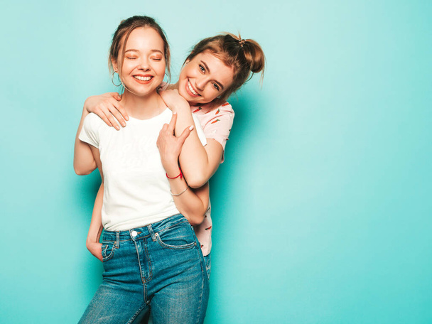 Deux jeunes belles filles hipster souriantes et blondes en jeans tendance d'été. Sexy femmes insouciantes posant près du mur bleu. Modèles à la mode et positifs s'amuser - Photo, image