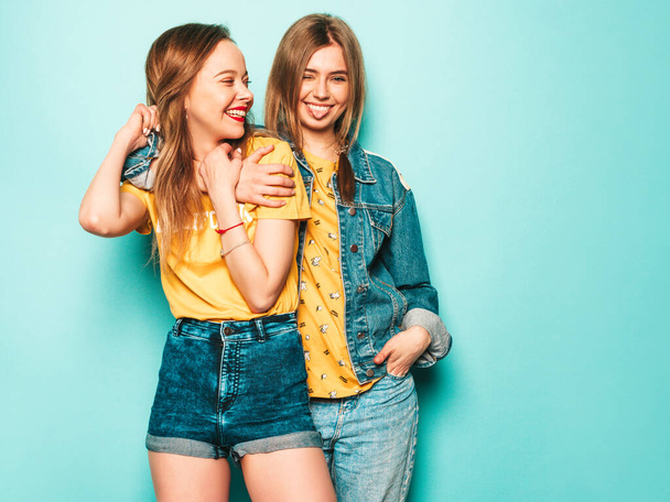 Two young beautiful smiling hipster girls in trendy summer yellow T-shirts and jeans jacket. Sexy carefree women posing near blue wall. Trendy and positive models show tongue - Фото, зображення