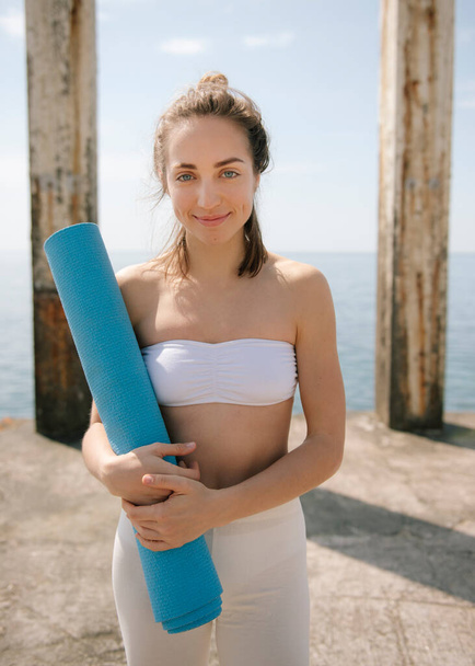 Smiling young woman holding yoga mat while standing on pier at seaside - Fotoğraf, Görsel