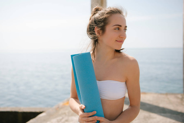 Smiling young woman holding yoga mat while standing on pier at seaside - Фото, изображение