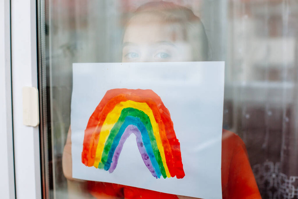 Маленькая девочка смотрит на бесконечность через окно и показывает фотографию радуги во время карантина коронавируса
 - Фото, изображение
