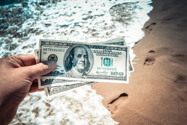 Girl holding money bill of 300 dollars on background of sea ocean waves with white foam and sand wet beach with footprints in sand close-up. Concept finance money holiday traveling dollars vacation - Foto, afbeelding