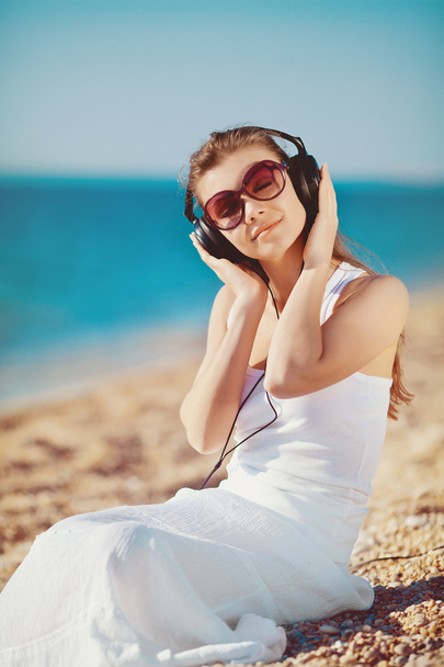 Beautiful woman listening to music against blue sky in headphones on the seashore - Fotó, kép