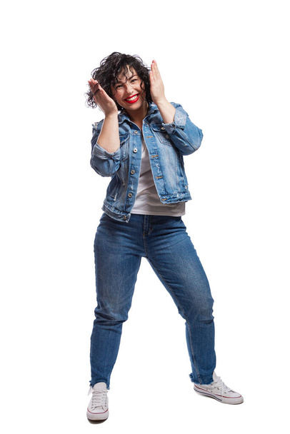Happy dancing young girl in jeans. Positive and happy. Bright brunette with curly hair and red lipstick. Isolated on a white background. Vertical. - 写真・画像