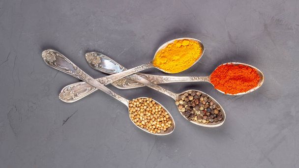 Turmeric powder, paprika, coriander and black peppercorns in metal teaspoons on a gray background - Photo, Image