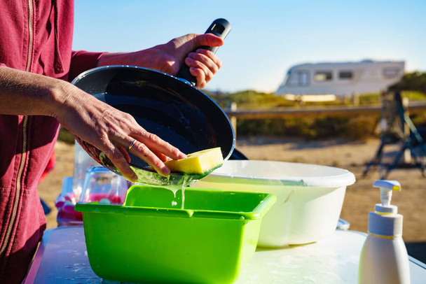 Mature woman washing up dishes in bowl on fresh air. Dishwashing outdoor on camping site, sea shore - Fotoğraf, Görsel