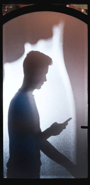 Fiatal fiú sziluett áll az üvegajtó mögött, nézi a mobilját. - Fotó, kép