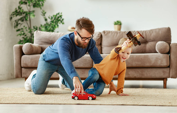 Seitenansicht eines fröhlichen jungen Mannes und kleinen Jungen in lässiger Kleidung, die auf dem Boden sitzen und mit Spielzeugauto und Flugzeug spielen, während sie Zeit miteinander verbringen - Foto, Bild