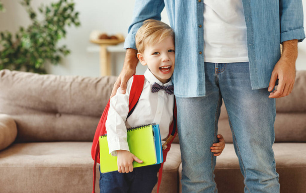 glücklich lachendes Kind ein kleiner Junge mit Schulrucksack und Büchern umarmt das Bein seines Vaters und macht sich bereit für das neue Schuljahr at hom - Foto, Bild