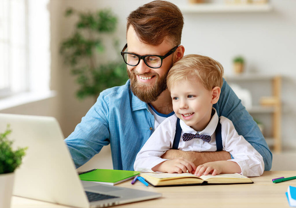 Молодий бородатий чоловік в окулярах вказує на ноутбук, пояснюючи урок маленькому синові під час онлайн-навчання разом вдома
 - Фото, зображення