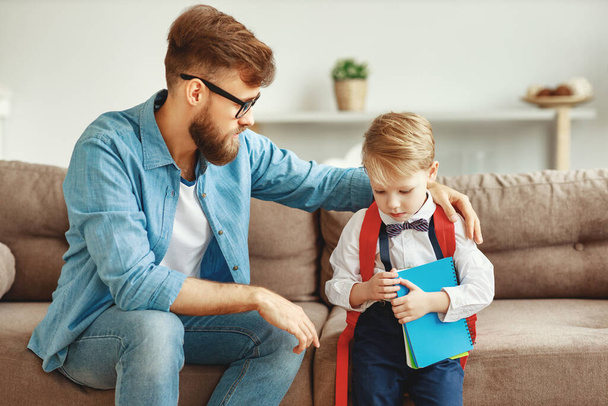 Любящий отец обнимает и поддерживает расстроенного маленького сына школьными вещами, сидя вместе на диване у дома
 - Фото, изображение