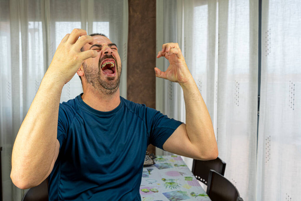 Γενειοφόρος άντρας με σκούρο μπλε πουκάμισο και έκφραση οργής στο σαλόνι του σπιτιού. Διασκεδαστική ιδέα - Φωτογραφία, εικόνα