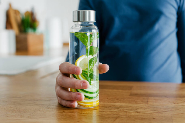 Teenager boy holding glass bottle to go infused detox water with cucumber, lemon and mint kitchen on background. Healthy drink for healthy life. Copy space - Photo, Image