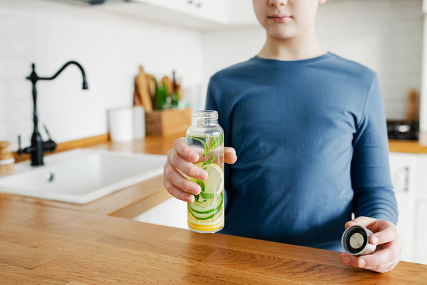 Teenager chlapec drží skleněné láhve naplněné detoxikační vodou s okurkou, citronem a mátovou kuchyní na pozadí. Dieta, zdravé stravování, koncepce hubnutí. Kopírovat prostor - Fotografie, Obrázek
