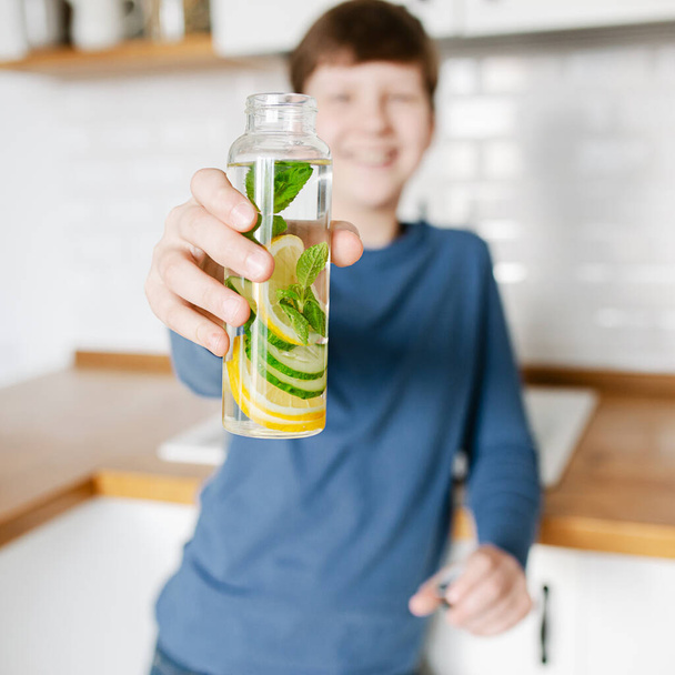 Teenager boy holding glass bottle infused detox water with cucumber, lemon and mint kitchen on background. Diet, healthy eating, weight loss concept. Copy space - Photo, Image