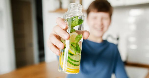 Teenager boy holding glass bottle infused detox water with cucumber, lemon and mint kitchen on background. Healthy drink for healthy life. Copy space - Photo, Image