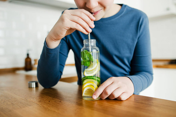 Teenager boy drinking infused detox water with cucumber, lemon and mint from glass bottle using stainless steel straw kitchen on background. Healthy drink for healthy life. Copy space - Photo, Image