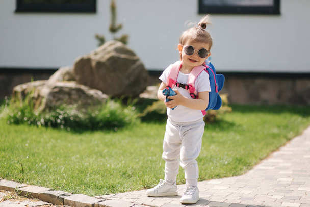 Szczęśliwa mała dziewczynka wchodzi do domu z plecakiem. Cute półtora roku dziewczyna w okularach przeciwsłonecznych - Zdjęcie, obraz