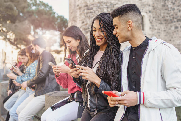 Group of generation z young people chatting together sitting on a bench outdoors. Students having fun together. Focus on a mixed race girl smiling and using social media on her smartphone - Fotoğraf, Görsel