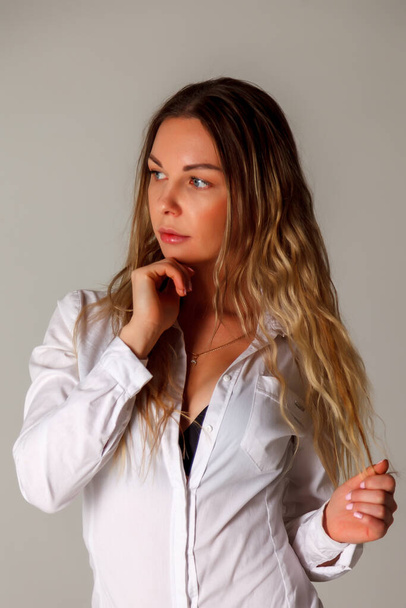 Girl of Slavic appearance in white shirt on gray background Young woman sits with positive gesture of hands dressed in homemade shirt showing beauty posing An empty copy of space for your ad or text - Foto, afbeelding