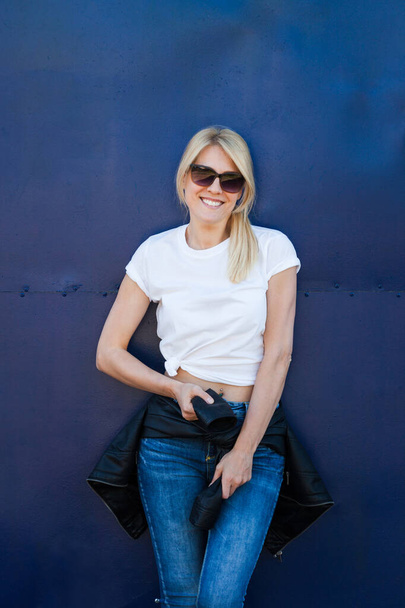 junge blonde lächelnde Frau in lässigem weißem T-Shirt und Jeans mit Sonnenbrille vor blauem Metallhintergrund in T-Shirt-Attrappe - Foto, Bild