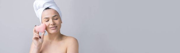 Krásný portrét ženy v bílém ručníku na hlavě s houbičkou pro tělo s výhledem na růžové srdce. Péče o pleť čištění ekologické kosmetické lázně relaxační koncept. - Fotografie, Obrázek