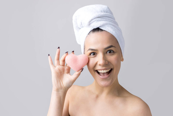Krásný portrét ženy v bílém ručníku na hlavě s houbičkou pro tělo s výhledem na růžové srdce. Péče o pleť čištění ekologické kosmetické lázně relaxační koncept. - Fotografie, Obrázek