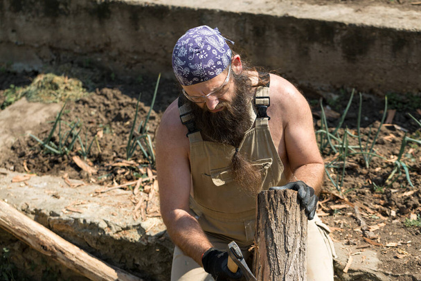 Um homem barbudo de meia-idade numa bandana corta troncos com um machado. Brutal em macacão faz o trabalho duro em um dia quente
. - Foto, Imagem