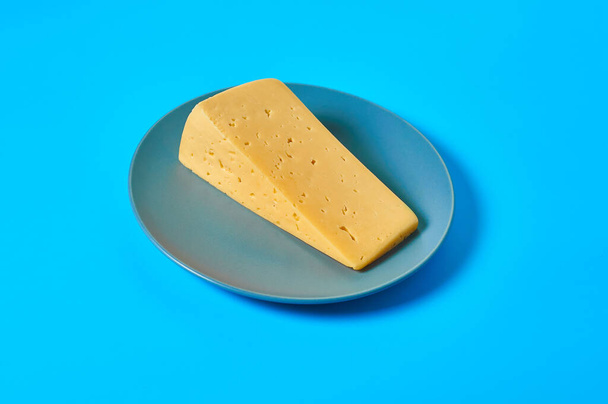 One piece of cheese in form of triangle on gray round plate lies on blue desk on kitchen or market. Close-up - Foto, Imagem