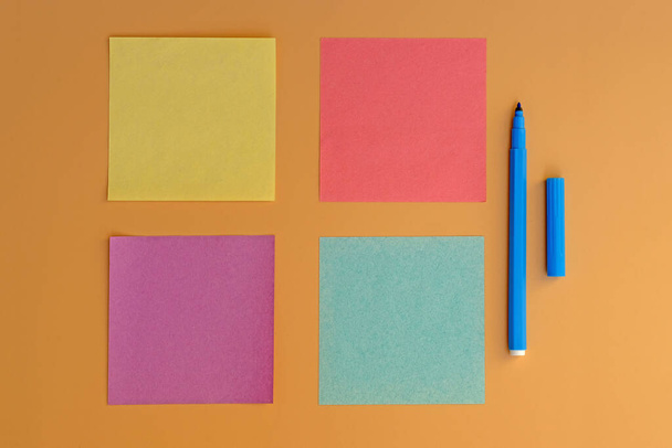 Φυλλάδια διαφόρων χρωμάτων για σημειώσεις και ένα μπλε μαρκαδόρο για γραφή. Αυτοκόλλητα κίτρινου, πράσινου, ροζ χρώματος top view. Κενό μικρό τετράγωνο χαρτί σημειώσεις σε ένα μπεζ φόντο. - Φωτογραφία, εικόνα